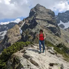 Kobieta z kijkami trekkingowymi na skalistej ścieżce w Dolinie Gąsienicowej. W tle majestatyczny szczyt i ośnieżone zbocza.