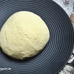 Frischer pizzateig auf schwarzer Platte, bereit für den pizzaofen. Ein Holzschaber liegt daneben.