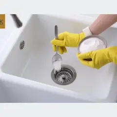Ręce w żółtych rękawiczkach wsypują sodę do odpływu zlewu, by zapobiec zapowietrzonym rur odpływowym.