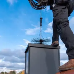 Ein Schornsteinfeger steht auf einem Dach und lässt eine Bürste in den Schornstein von unten kehren.