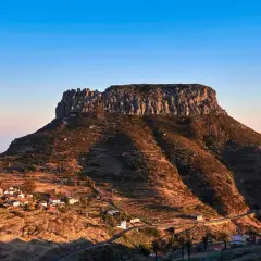 La imponente fortaleza de Chipude se alza sobre un paisaje rural con casas blancas y palmeras, bajo un cielo azul claro.