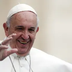 Papież Franciszek w białej sutannie i piusce, uśmiechnięty, macha ręką.