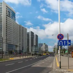 Ulica z nowoczesnymi budynkami i tramwajem. Na słupie widoczny znak D3 informujący o zakazie zatrzymywania się i dozwolonych kierunkach jazdy.