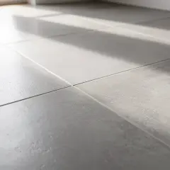 Lumière du soleil traversant un carrelage gris, créant des ombres douces. On entendrait presque le carrelage qui sonne creux sous les pas.