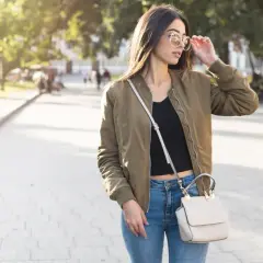 Młoda kobieta w stylowej kurtce wiosennej khaki, jeansach i z torebką na ramieniu spaceruje po parku.