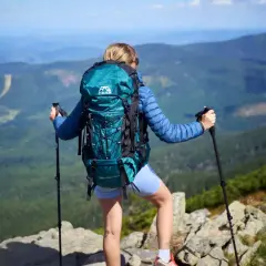 Kobieta z turkusowym plecakiem i kijkami trekkingowymi podziwia widok z góry. Idealny plecak turystyczny na wyprawę.