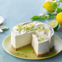 En krämig citron cheesecake med rivet citronskal som dekoration. Bredvid ligger färska citroner och en lime.