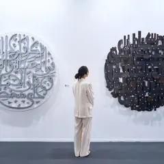 En kvinna i ljus kostym betraktar två runda konstverk med arabisk kalligrafi. Det vänstra är i metall, det högra i mörkare material. En atra daniel.