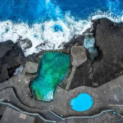 Piscinas naturales de agua turquesa y azul brillante, rodeadas de rocas volcánicas y el mar embravecido.