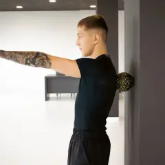 Mężczyzna z tatuażem na ramieniu masuje plecy piłką, celując w punkty spustowe.