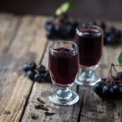 Dwa kieliszki z ciemnym płynem, obok kiście aronii i goździki. Szybka nalewka z aronii gotowa do degustacji.