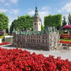Miniatura ratusza z wieżą zegarową, otoczona czerwonymi kwiatami. Idealne miejsce na trasie po Kotlinie Kłodzkiej.