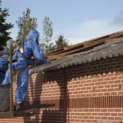 Dwóch pracowników w niebieskich kombinezonach ochronnych usuwa eternit z dachu. Azbest jest niebezpieczny.