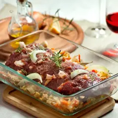 Surowe mięso z jelenia w naczyniu żaroodpornym, doprawione ziołami i cebulą, gotowe do pieczenia. Obok kieliszek czerwonego wina.