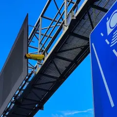 Żółta kamera na konstrukcji nad drogą, obok niebieskiego znaku z symbolami aparatów. To zapowiedź nowych odcinkowych pomiarów prędkości.