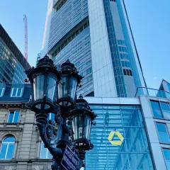Ein historischer Laternenpfahl vor dem modernen Commerzbank-Gebäude in Frankfurt. Die Fassade spiegelt den blauen Himmel wider.