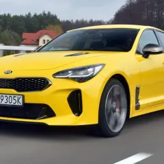 Żółty Kia Stinger GT pędzi po drodze, otoczony zielenią i domami.