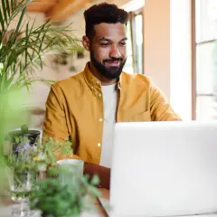 Uomo sorridente al laptop, circondato da piante. Sembra che stia imparando nuovi modi di dire per arricchire la sua comunicazione.