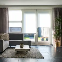 Grå soffa i vardagsrummet med draperade gardiner och stora fönster som släpper in ljus.