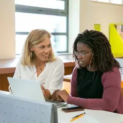 Dos mujeres en un aula, una rubia y otra afrodescendiente, revisan un portátil. La rubia sonríe, la otra parece preocupada, quizás porque su aptis caduca.