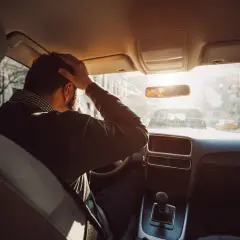 Hombre con mano en la cabeza dentro de un coche, buscando trucos para conducir sin nervios. El sol brilla a través del parabrisas.