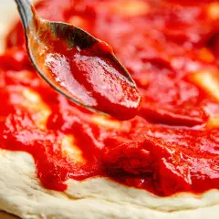 Eine Löffelspitze verteilt die leuchtend rote Pizza Sauce wie in der Pizzeria auf den Teig.