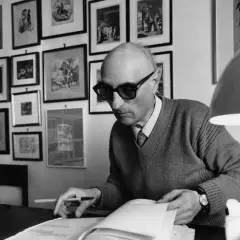 Gesualdo Bufalino, con occhiali scuri, è immerso nella lettura di un libro, circondato da quadri alle pareti.
