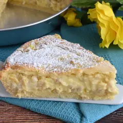 Una fetta di pastiera napoletana con crema pasticcera, cosparsa di zucchero a velo, su un piatto bianco, con fiori gialli sullo sfondo.