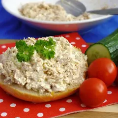 Pyszna pasta z tuńczyka i twarogu na bułce, ozdobiona natką pietruszki, obok pomidorki i ogórek.