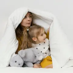 Mama i dziecko bawią się pod białą kołdrą, która przypomina przytulne kołderki dla niemowląt. Obok leży szary pluszowy króliczek.