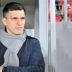 Robert Podoliński, trener w szaliku, spogląda w bok na tle stadionu z czerwonymi siedzeniami.