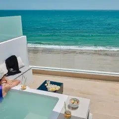 Mujer en bañera de hidromasaje con vista al mar, con presillas bajas en su traje de baño, disfruta de una bebida refrescante.