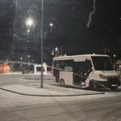 Biały bus na ośnieżonym przystanku nocą, ozdobionym świątecznymi lampkami.