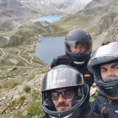 Tre motociclisti con caschi ammirano il panorama mozzafiato del Col del Nivolet, con i suoi laghi alpini e le strade tortuose.