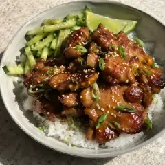 En skål med ris, gurka och saftig grillad kyckling med sesamfrön och salladslök. En limeklyfta ligger bredvid.