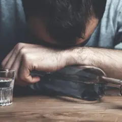 Zmęczony mężczyzna opiera głowę na butelce alkoholu, obok stoi kieliszek wody. To symboliczny obraz odstawienia alkoholu i walki o trzeźwość.
