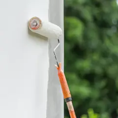 Fassade streichen ohne Reinigung: Ein Malerroller mit orangefarbenem Griff überzieht eine weiße Wand mit Farbe. Im Hintergrund verschwimmen grüne Bäume.