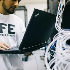 Inżynier w białej koszulce z napisem "FE FIELD ENGINEER" sprawdza połączenia sieciowe na laptopie, analizując ruch icmp.