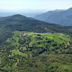 Vista aérea del **santuari del far**, rodeado de verdes colinas y bosques frondosos. Caminos serpentean entre prados y edificios dispersos.