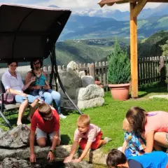 Famiglia rilassata in un giardino con vista sulle montagne, tipico delle fattorie altoatesine. Bambini giocano vicino a un laghetto, mentre adulti riposano su un'altalena.