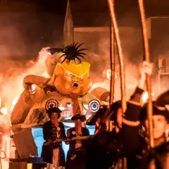 Un pupazzo gigante con un cappello giallo, circondato da fiamme e persone in costume, celebra un giorno festivo in inglese.
