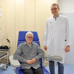 Ein Arzt steht neben einem Patienten, der für eine Alzheimer Behandlung in einem Behandlungsstuhl sitzt. Infusionsständer und medizinische Geräte sind im Hintergrund sichtbar.