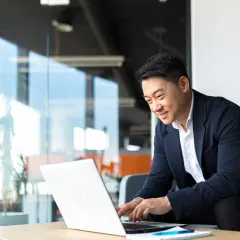 An Asian man in a suit smiles while working on his laptop, perhaps studying for his bachelors in organizational leadership.