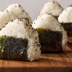 Several triangle sushi onigiri, sprinkled with sesame seeds and wrapped in nori, sit on a wooden board.