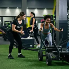 Intensywny trening interwałowy na siłowni. Kobieta pcha sanie, mężczyzna ćwiczy z linami, a inni wykonują ćwiczenia z kettlebell.