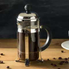 Szklany french press z ciemną kawą, obok biała filiżanka i ziarna kawy. Idealny poranek z kawą do french press.