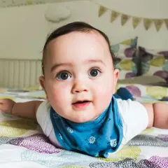 Un adorabile neonato di 4 mesi, sdraiato a pancia in giù sul letto, con gli occhi spalancati e un bavaglino blu.