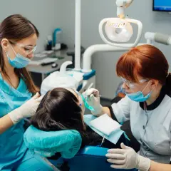 Dwie kobiety w maseczkach i okularach ochronnych pracują przy pacjencie w gabinecie stomatologicznym. Jedna z nich, asystentka stomatologiczna, podaje narzędzia.