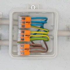 Nowoczesna instalacja elektryczna z kostkami WAGO 221, łączącymi przewody niebieski, żółto-zielony i brązowy w białej puszce.