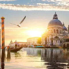 Góndola navega por canal veneciano al atardecer, con la Basílica de Santa Maria della Salute y otras **ciudades de Europa** reflejadas en el agua.